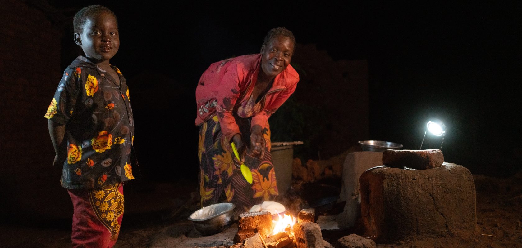 Kesilina and her son look to the camera while Kesilina cooks dinner using her solar light.