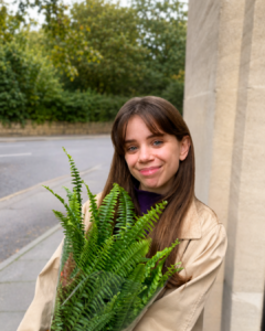 A smiling woman with long brown hair holds a lush green fern wrapped in clear plastic while standing beside a stone building on a quiet, tree-lined street.